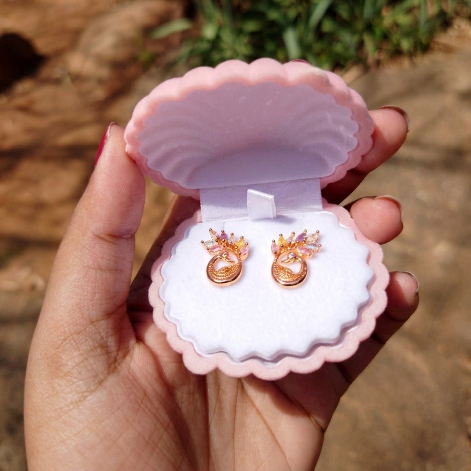 Pink shell-shaped jewelry box with gold earrings inside, held by a hand against a natural background.