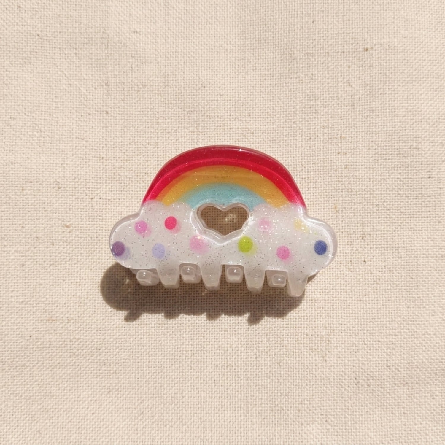 Hair clip with rainbow and cloud design on a beige fabric background